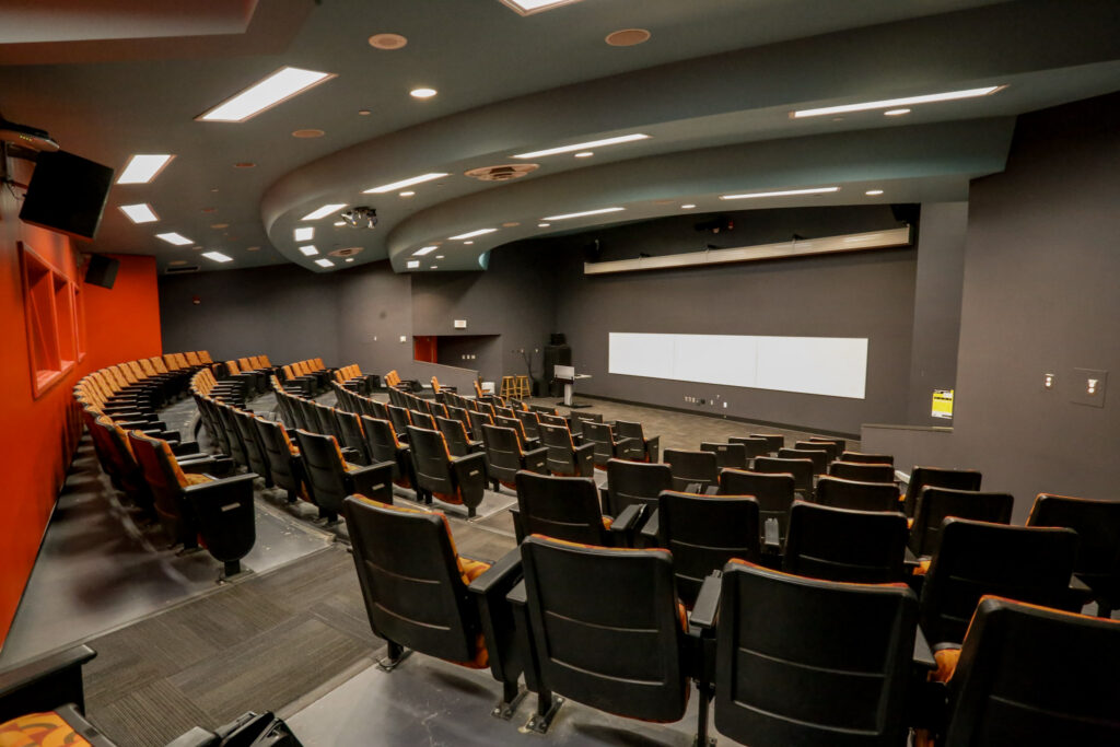 An empty, modern university lecture hall or auditorium with black and orange tiered seating, dark grey walls, a bright red accent wall, and a large white projection screen at the front.