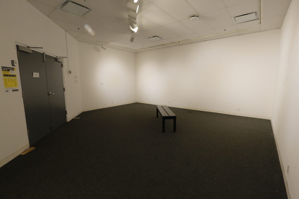 An empty art gallery or exhibition space with white walls, dark carpet, and a simple dark bench. Track lighting illuminates the walls, and a prominent emergency exit door with a yellow sign is on the left.