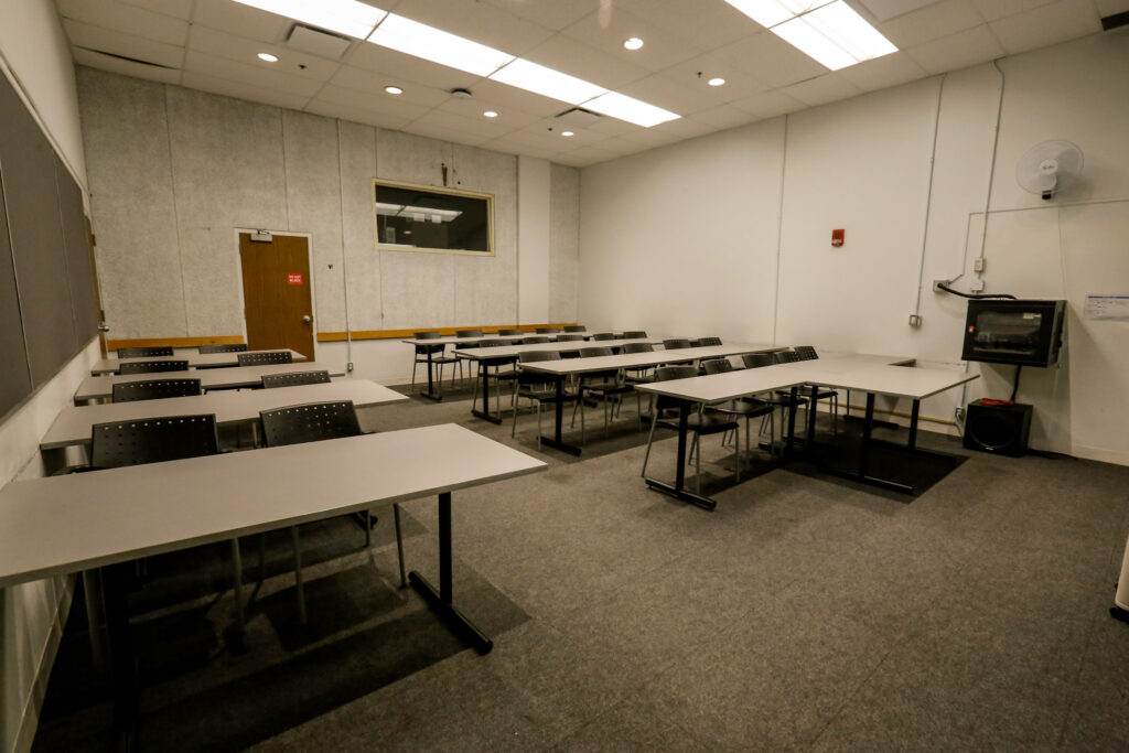 An unoccupied classroom or training room featuring modular grey tables and black chairs, with a door and wall-mounted electronics visible.