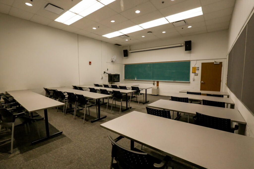 An empty, brightly lit modern classroom featuring rows of light gray tables and black chairs, a green chalkboard, and a projector screen.