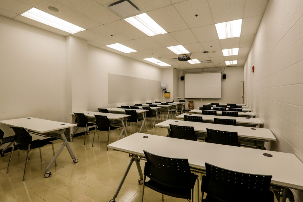 An unoccupied modern classroom or training room, featuring multiple rows of white tables and black chairs facing a projector screen and whiteboards.