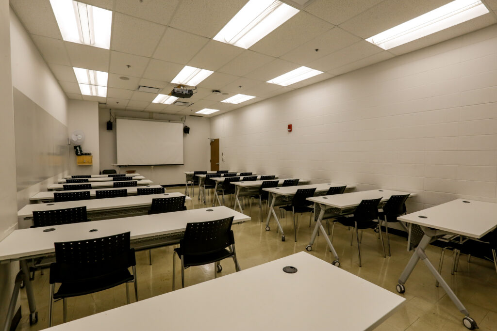 An empty, well-lit modern classroom featuring multiple rows of white tables and black chairs, arranged to face a large projection screen and a ceiling-mounted projector.