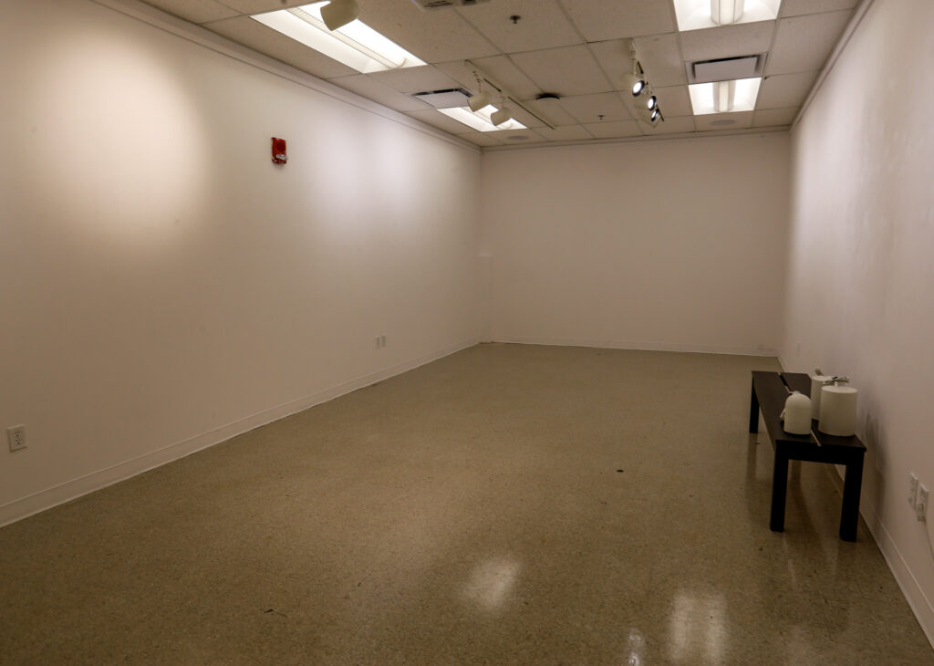 A well-lit, empty office or gallery room with white walls, a speckled tan floor, and a dark bench with two white decorative cylinders on the right. A red fire alarm is on the left wall.
