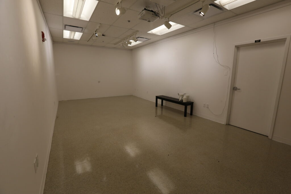 A modern, empty room or gallery space with white walls, a light brown floor, and track lighting, featuring a black bench with white objects and a projector.