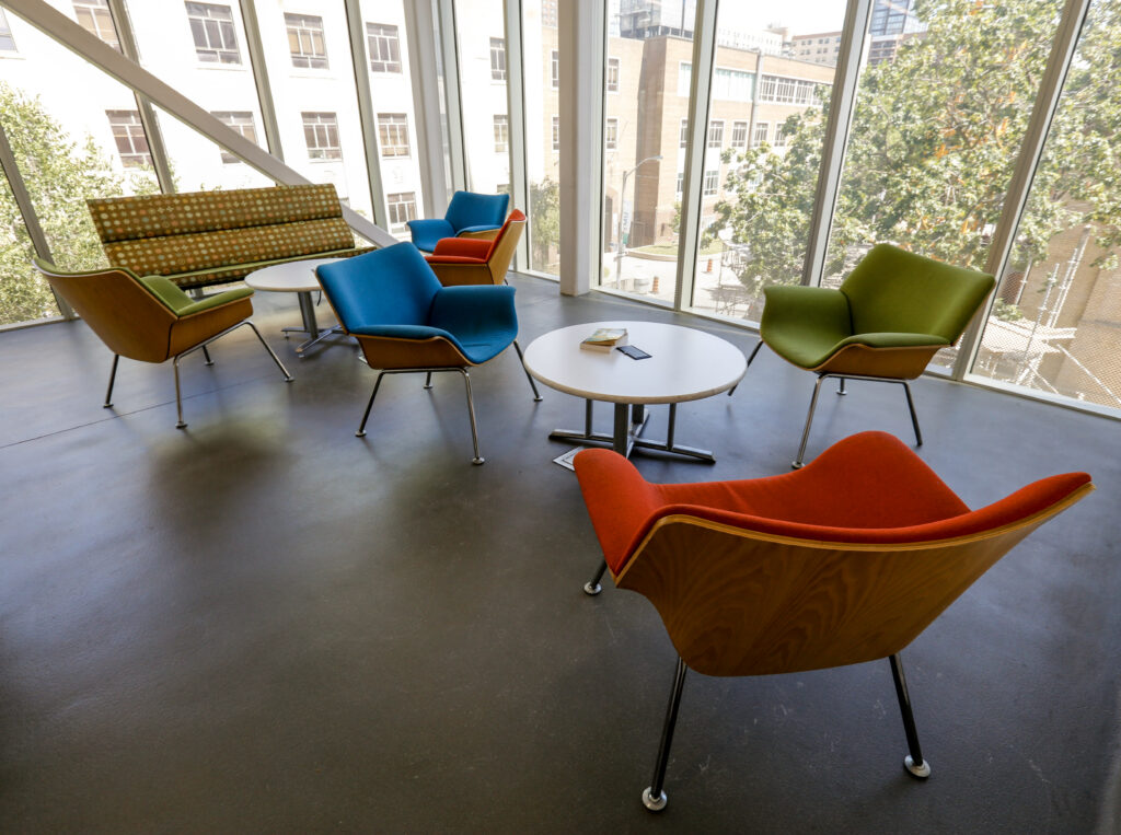 A bright, contemporary lounge featuring colorful mid-century modern style chairs in blue, green, and orange, a patterned green sofa, and white round tables, positioned alongside large windows offering an urban view.