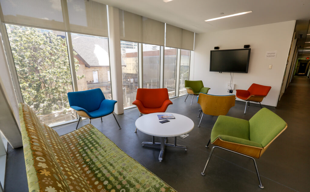 A bright, modern lounge or waiting area featuring a patterned sofa, several colorful mid-century style chairs, a coffee table, a wall-mounted TV, and large windows looking out at trees and buildings.