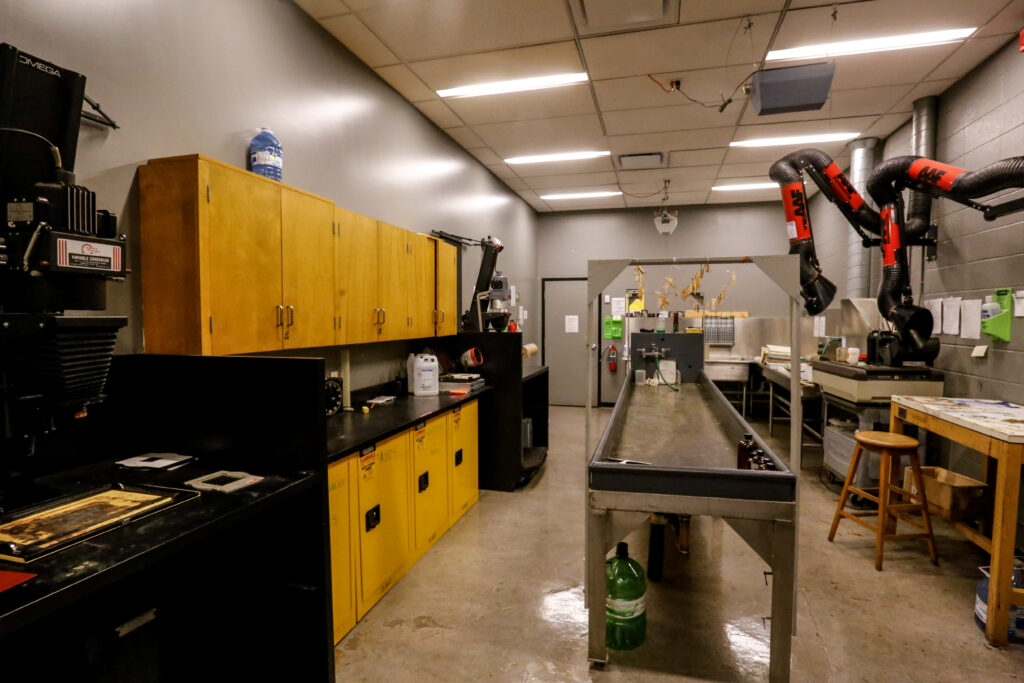 An interior shot of a professional darkroom featuring an Omega enlarger, yellow chemical storage cabinets, a long wet processing sink with items drying overhead, and prominent red ventilation arms.