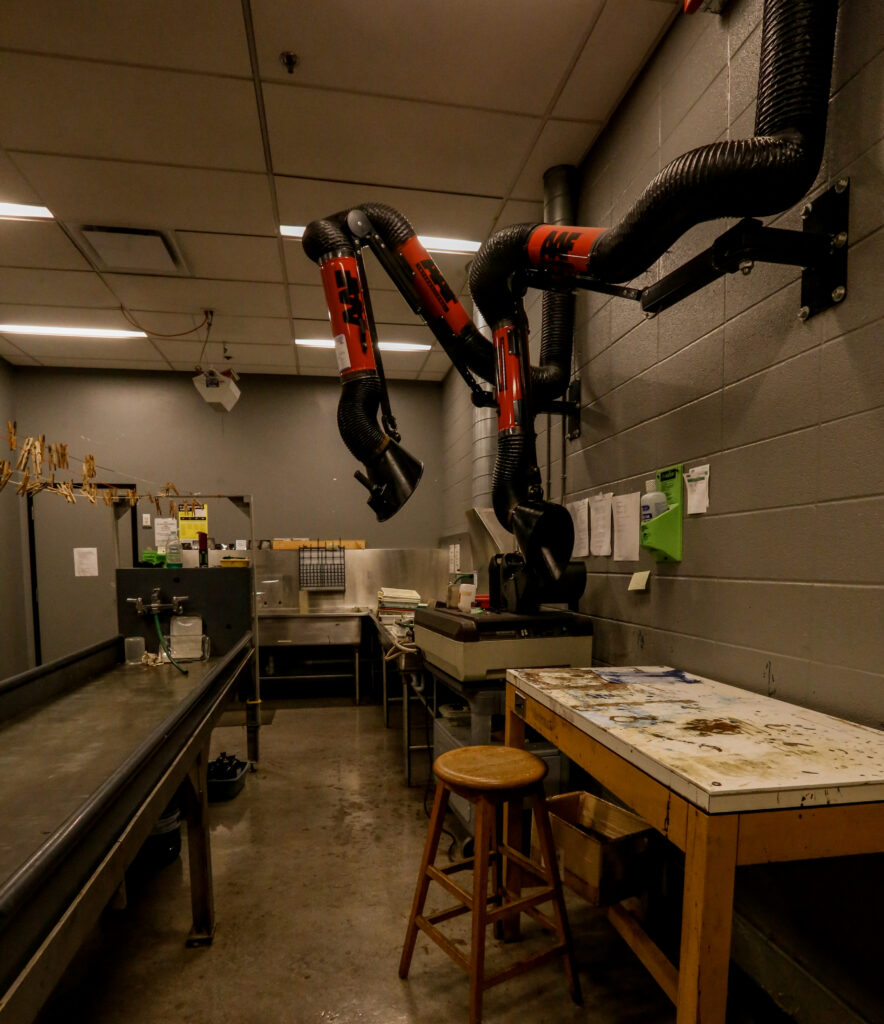 An untidy photo lab or art studio, featuring long stainless steel sinks, multiple red and black chemical fume extractor arms mounted on gray block walls, and a heavily stained wooden workbench in the foreground.