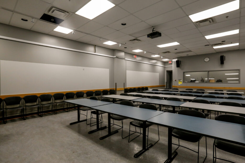 A well-lit, unoccupied modern classroom or training room, featuring rows of blue-topped tables with black chairs, a whiteboard wall, and a ceiling-mounted projector. An observation window is visible on the right wall.
