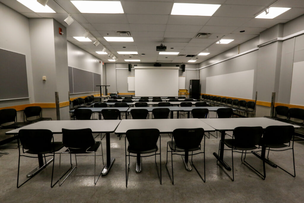 An empty, brightly lit classroom or training room with rows of black chairs and light gray tables facing a projection screen and dark gray wall panels.