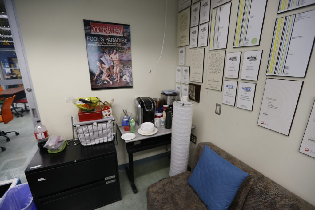 A common area in a journalism office featuring a Keurig coffee station, a wall of national magazine awards, a 'Fool's Paradise' journalism poster, and a patterned sofa.