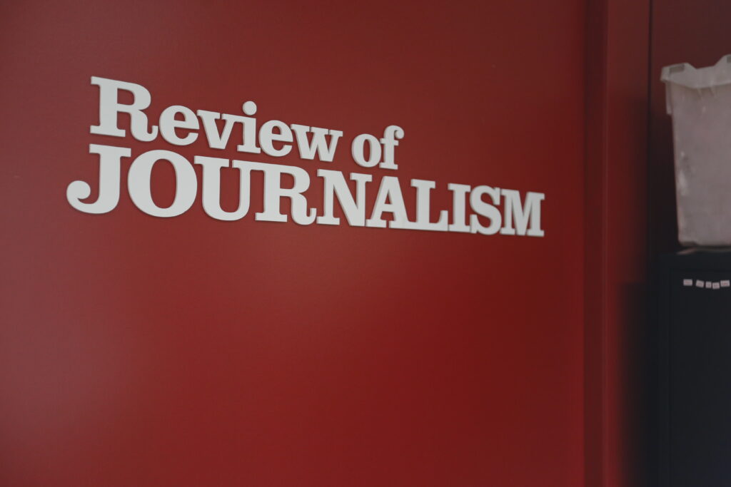 3D white letters spelling 'Review of JOURNALISM' mounted on a vibrant red wall, with a partial view of a dispenser to the right.