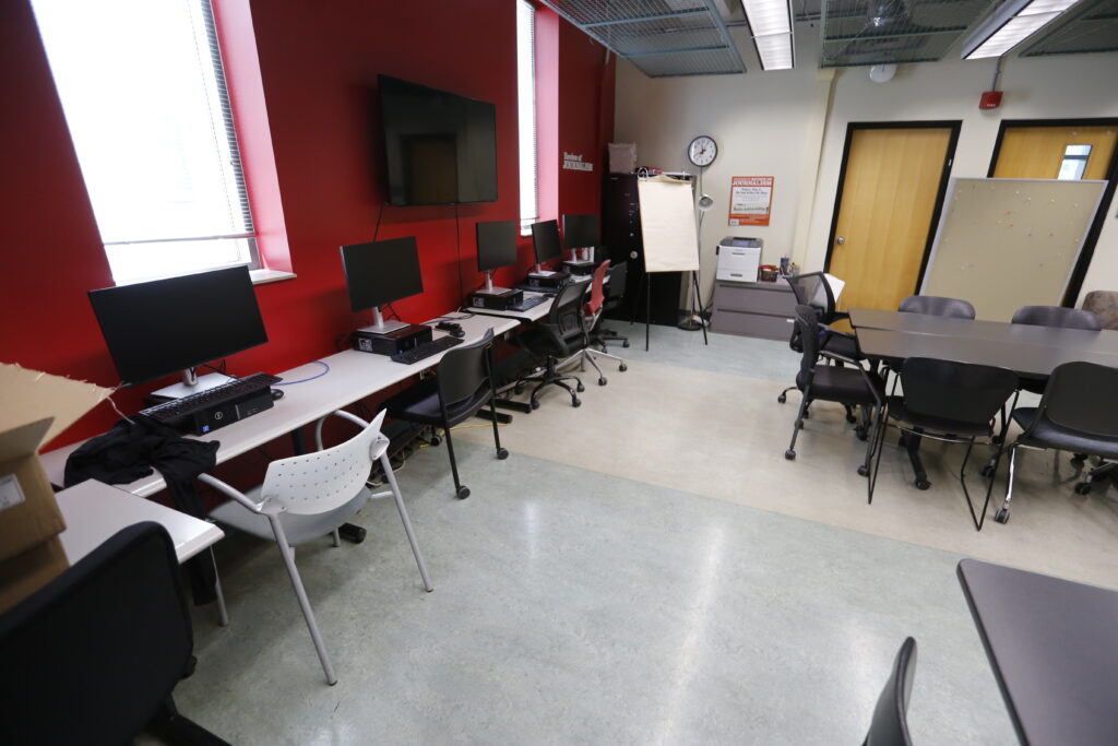 An empty modern classroom or journalism lab, featuring computer workstations along a red wall and a central conference table.