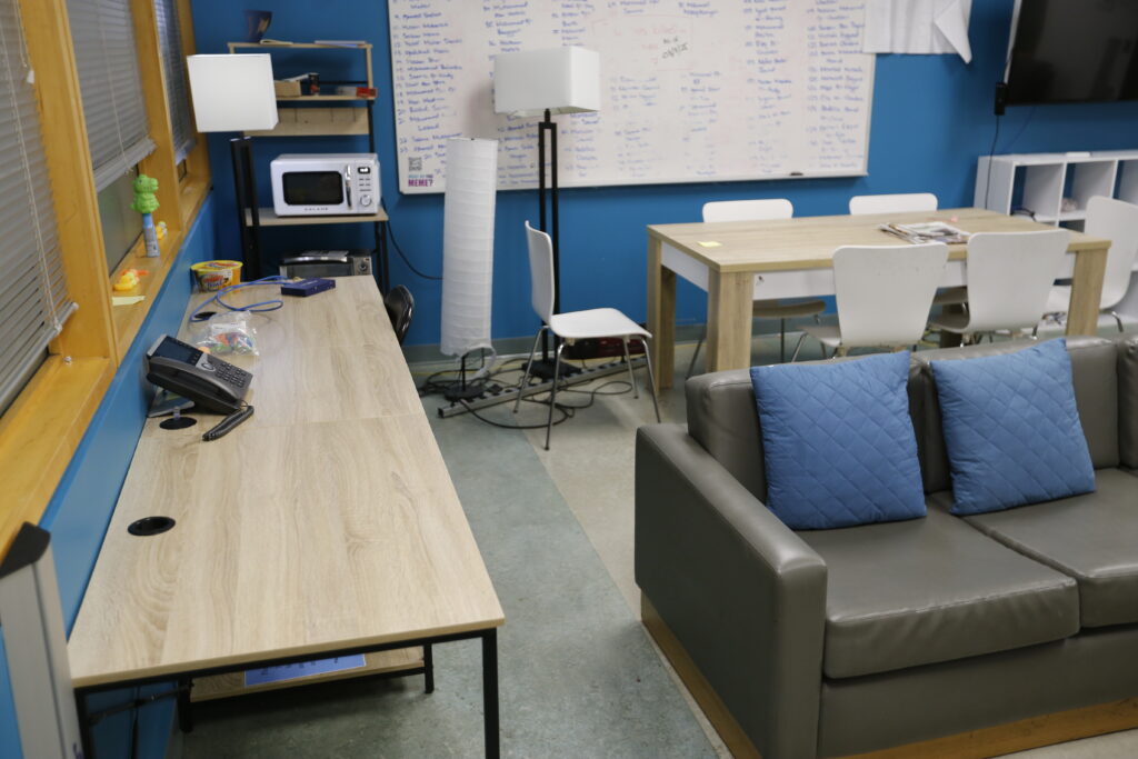 An informal office common area featuring a long desk by a window, a central dining table with white chairs, and a grey couch, all set against a vibrant blue wall with a whiteboard.