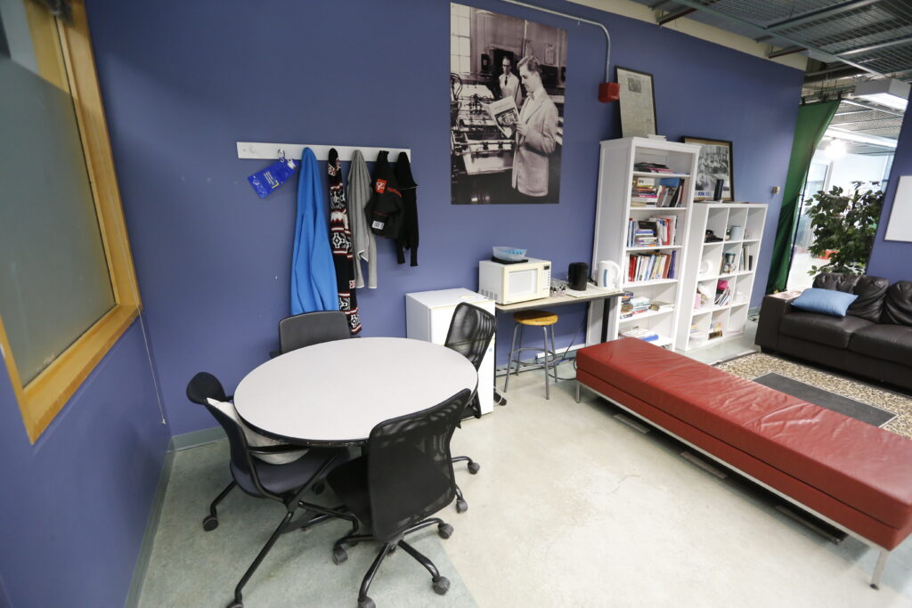 A casual common room with periwinkle blue walls, featuring a round table, a small kitchenette, white bookshelves, a long red bench, a brown leather sofa, and a large vintage lab photo.