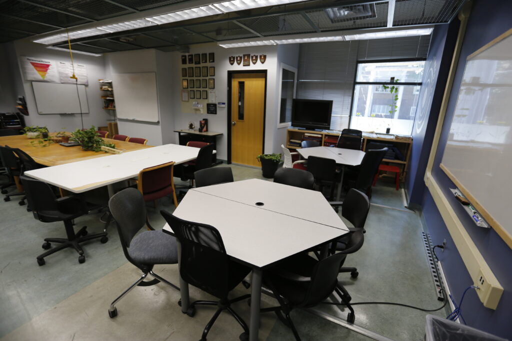 A well-lit, unoccupied classroom or conference room featuring multiple table configurations, including hexagonal and rectangular, surrounded by office chairs. Walls are adorned with whiteboards, informational posters, and framed awards.