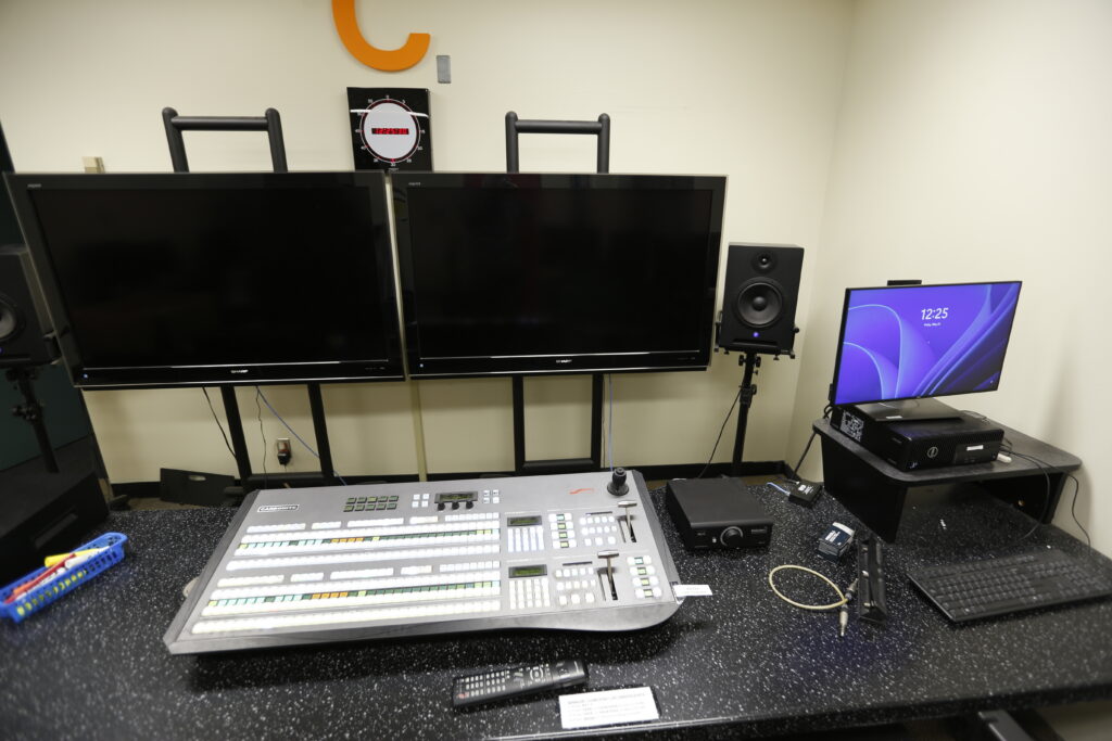A compact media control room setup featuring a large Ross Carbonite video switcher, two large off-screen monitors, a live clock, and a smaller computer monitor displaying the time.