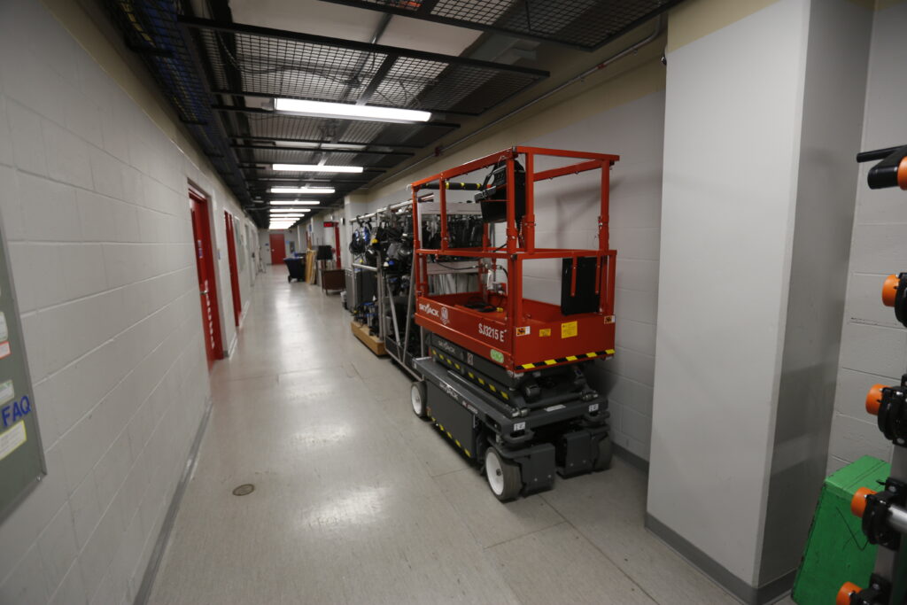 A red Skyjack scissor lift and equipment are stored along the wall of a modern, institutional hallway with exposed utilities overhead.