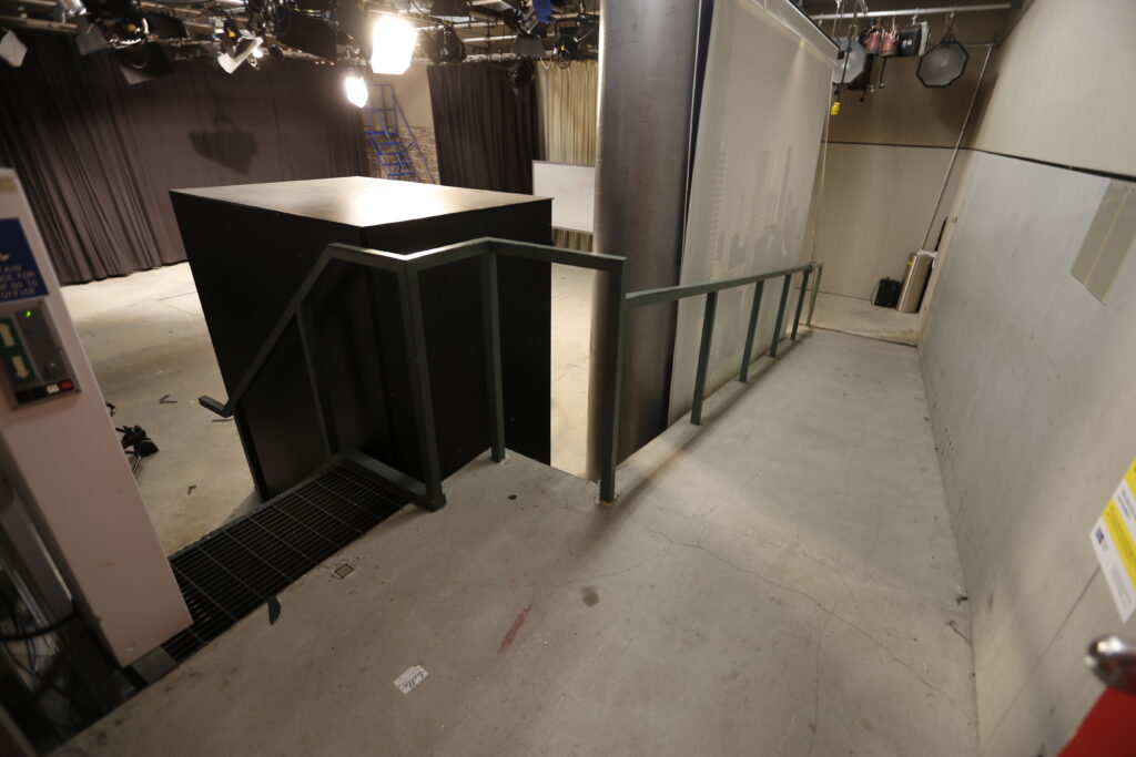 An interior shot of a television studio showing a concrete floor, a dark green railing, a black set piece, and overhead lights, with various backdrops including dark curtains and a cityscape.