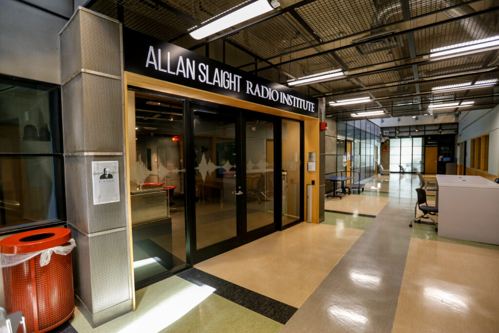 The entrance to the Allan Slaight Radio Institute, showing glass doors with sound wave decals, a plaque about Allan Slaight, and a brightly lit hallway stretching into the distance.