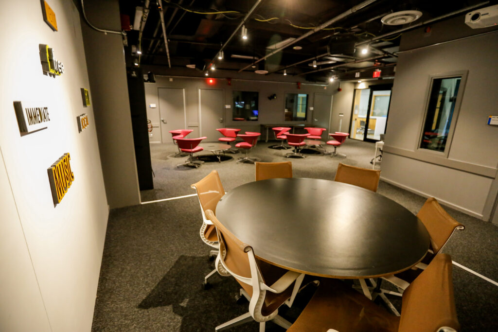 A contemporary collaborative workspace featuring a large oval table with brown chairs in the foreground, bright pink swivel chairs in the background, and 3D word art on the wall.
