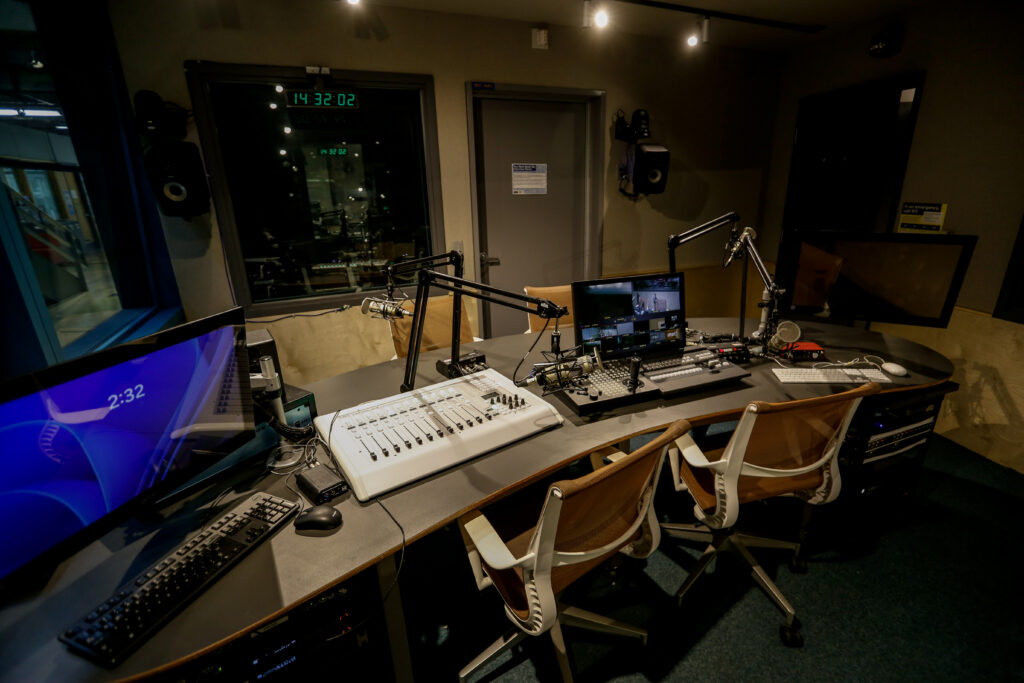 An empty professional broadcast studio control room features a curved desk with two microphones, an audio mixer, and video switching gear, facing multiple screens, one showing live feeds, another a digital clock at 14:32:02.