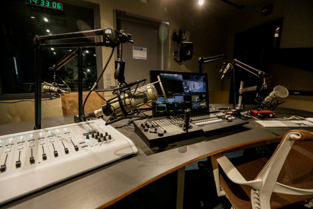 An illuminated broadcast studio control room features a desk with multiple microphones, a digital audio mixer, and a video switcher with a multiviewer monitor, indicating a live production setup.