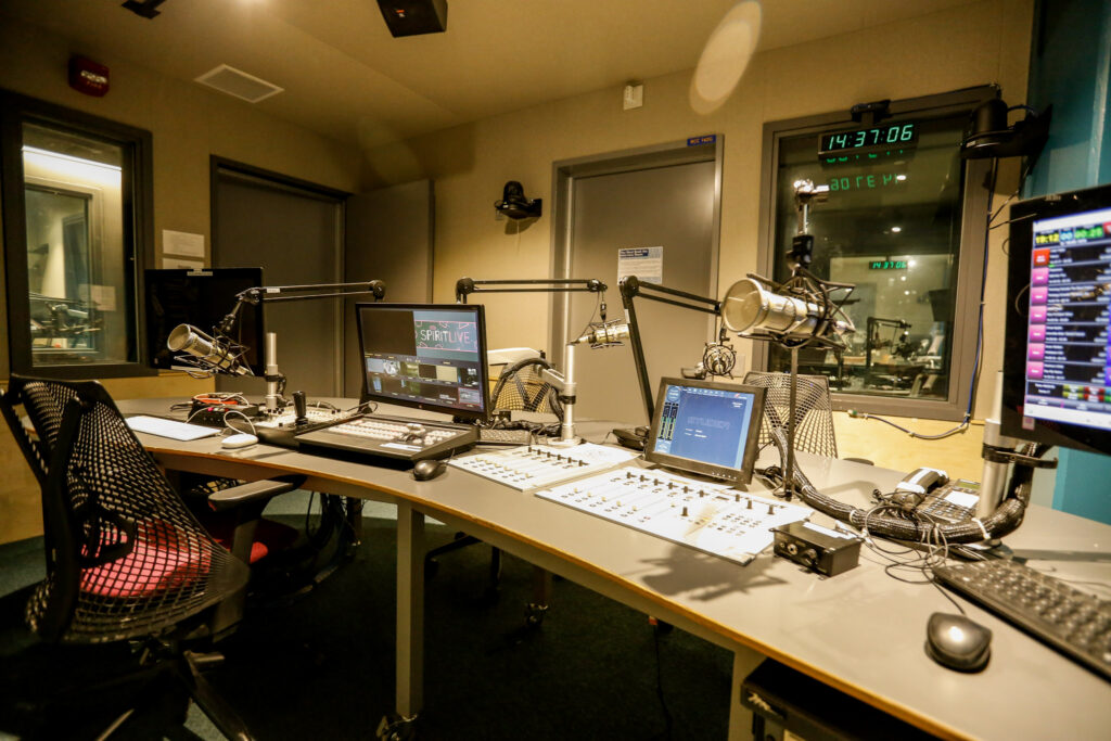 An empty radio studio with multiple microphones, computer monitors, and audio mixing equipment on a curved desk, overlooking an adjacent room through a large window.