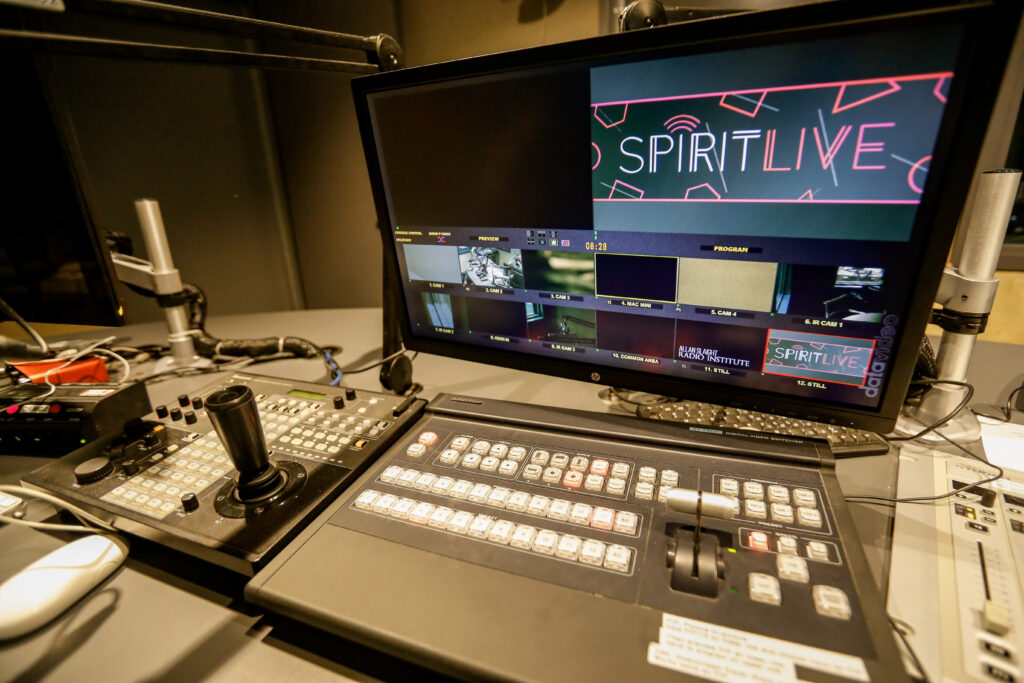 A broadcast control station featuring a multi-view monitor showing 'SPIRIT LIVE' graphics, live camera feeds, and 'Allan Slaight Radio Institute' text, alongside a camera joystick controller and a digital video switcher.