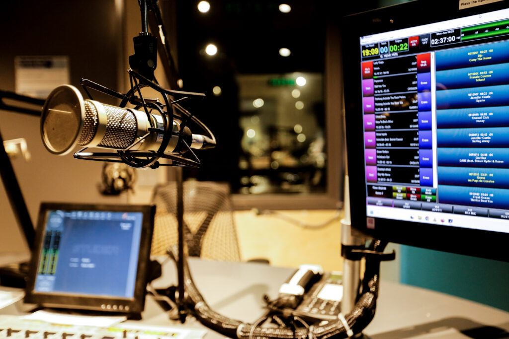A radio or podcast studio setup featuring a large microphone in the foreground, with two computer monitors displaying a playlist and audio controls in the background.