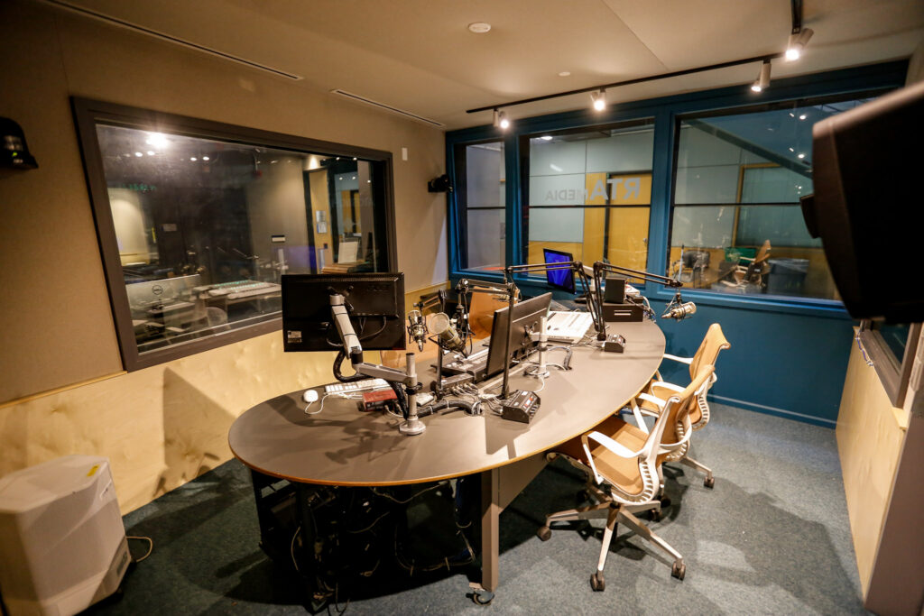 An empty, professional radio broadcast studio featuring a large oval desk with multiple microphones, monitors, and mixing consoles, with soundproof windows revealing a control room and a branded lounge area with 'RTR MEDIA' signage.