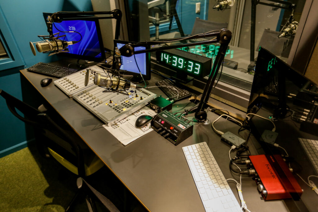 A professional radio broadcast studio workstation featuring two microphones on boom arms, an audio mixing console, computer monitors, and a prominent digital clock displaying 14:39:33.