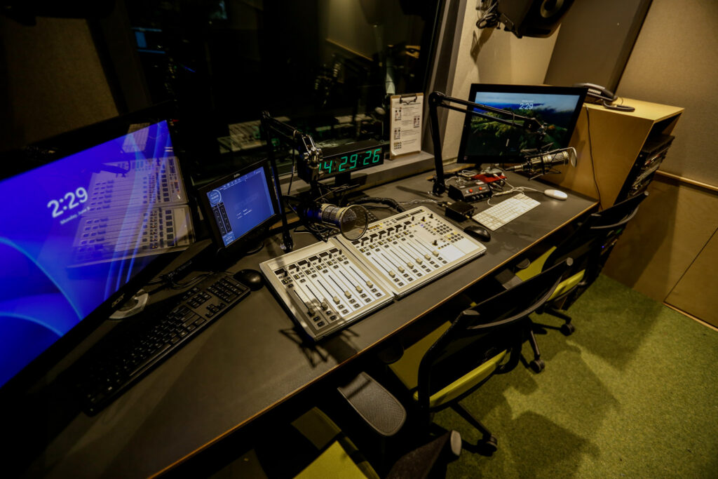 An empty broadcast studio setup with a long desk featuring two audio mixing consoles, multiple computer screens displaying clocks and audio software, and boom-mounted microphones.