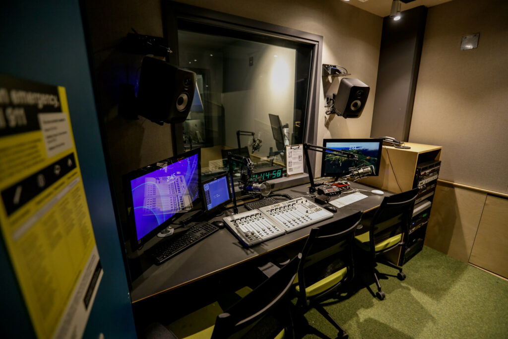 An empty, professional radio broadcasting or podcasting studio control room, featuring a large audio mixer, multiple computer screens, studio monitors, and a soundproof window looking into a separate recording space.