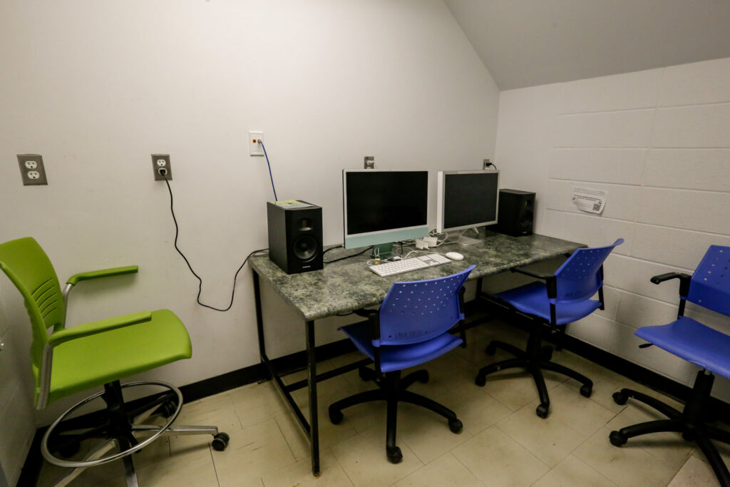 A small media or computer lab room with a long desk featuring two monitors, studio speakers, and three chairs (one green, two blue), against white walls.