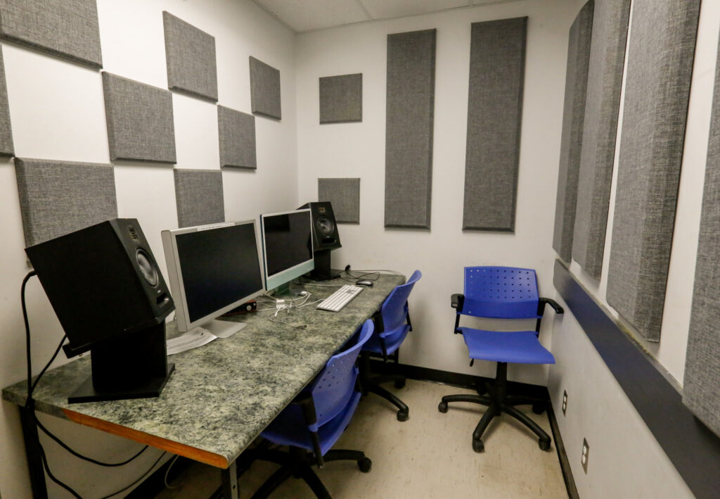 An audio or video editing suite featuring grey sound-absorbing panels on white walls, a long desk with multiple computer monitors and studio speakers, and three blue rolling chairs.
