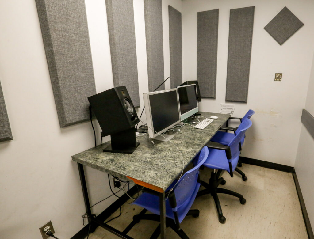 A small studio space with a desk, two computers, a large studio monitor, and three blue chairs, featuring grey acoustic panels on white walls.