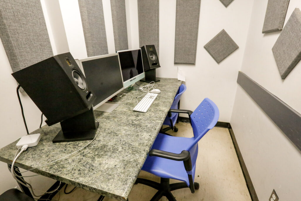 A small sound-treated audio editing workstation featuring a desk with two computer monitors, two studio speakers, and two blue chairs, surrounded by gray acoustic wall panels.