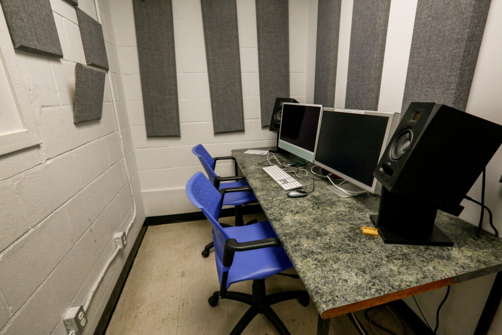 An equipped audio or video editing room with two computer monitors, studio speakers, and blue chairs, featuring acoustic panels on white cinder block walls.