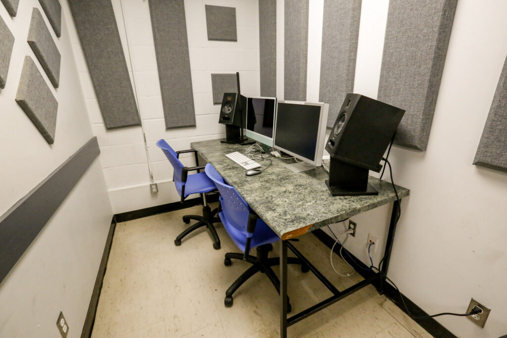 An acoustically treated studio booth featuring a granite-like desk with two computer workstations, two studio monitor speakers, and two blue chairs.