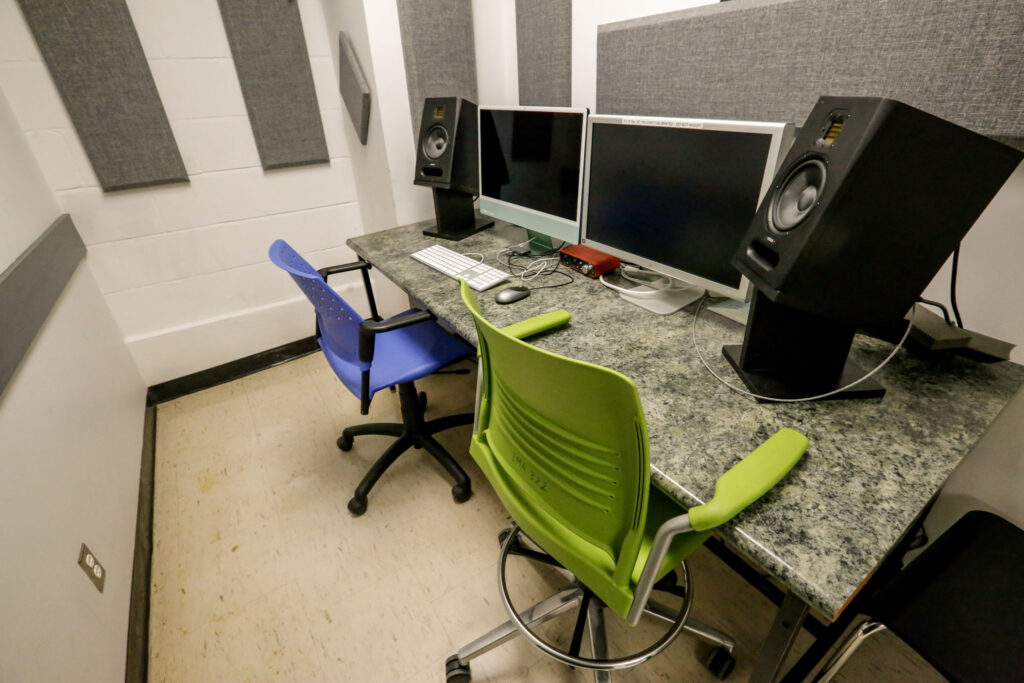 A small, sound-treated audio editing workstation features a desk with dual computer monitors, two studio speakers, a keyboard, and mouse. Two chairs, one blue and one lime green, are placed at the desk, with grey acoustic panels on the white walls.