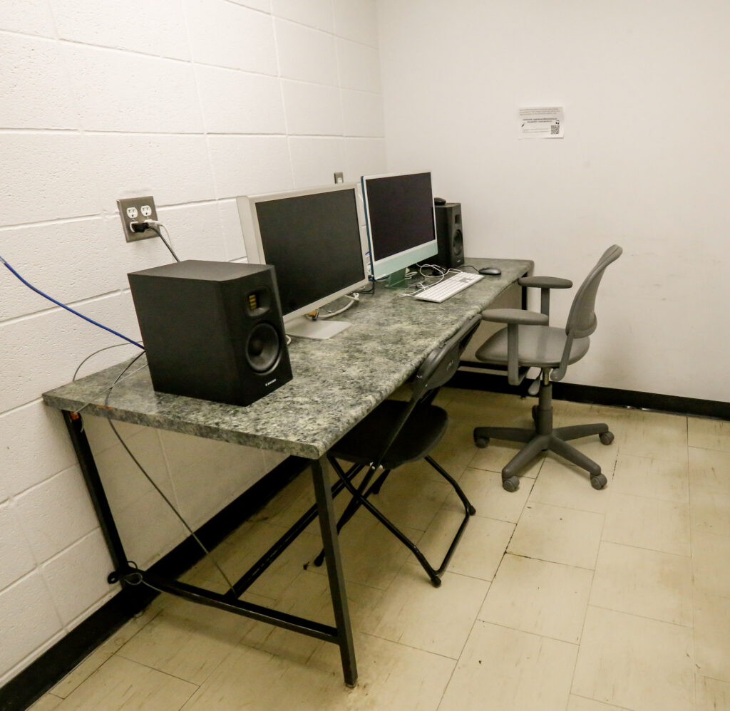 A desk with a speckled gray countertop holds two computer monitors, studio speakers, keyboard, and mouse, with two chairs in a room with white block walls and a tiled floor.
