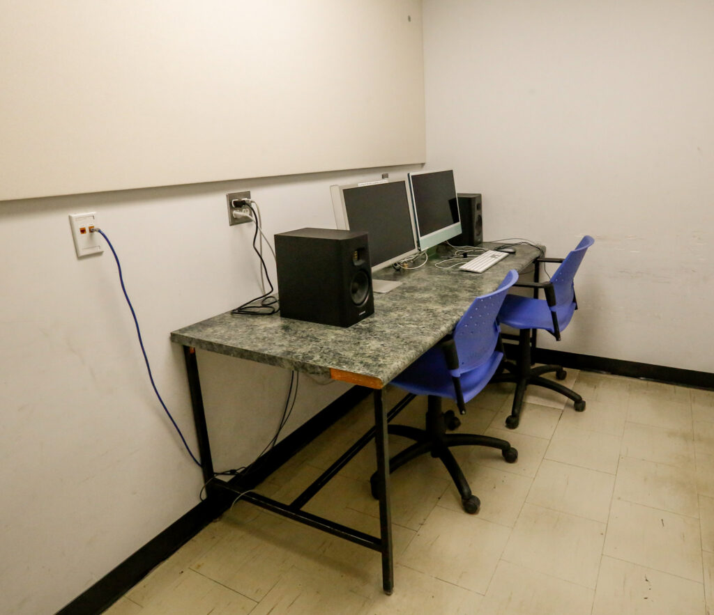 A dual computer setup with monitors, speakers, and blue office chairs on a granite-patterned desk in a room with white walls.