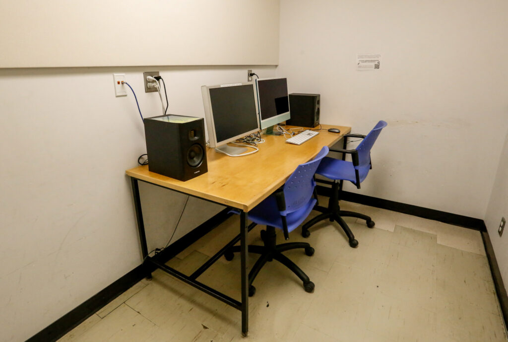 An audio editing workstation featuring two computers, two studio monitors, and two blue chairs, set up at a wooden desk in a simple lab or study room.