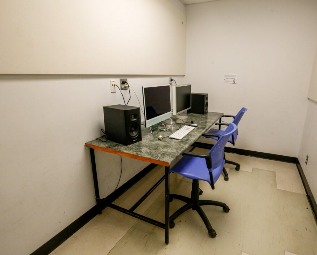A small computer lab or media editing station with two setups, each featuring a monitor, speakers, and a blue rolling chair at a shared desk.