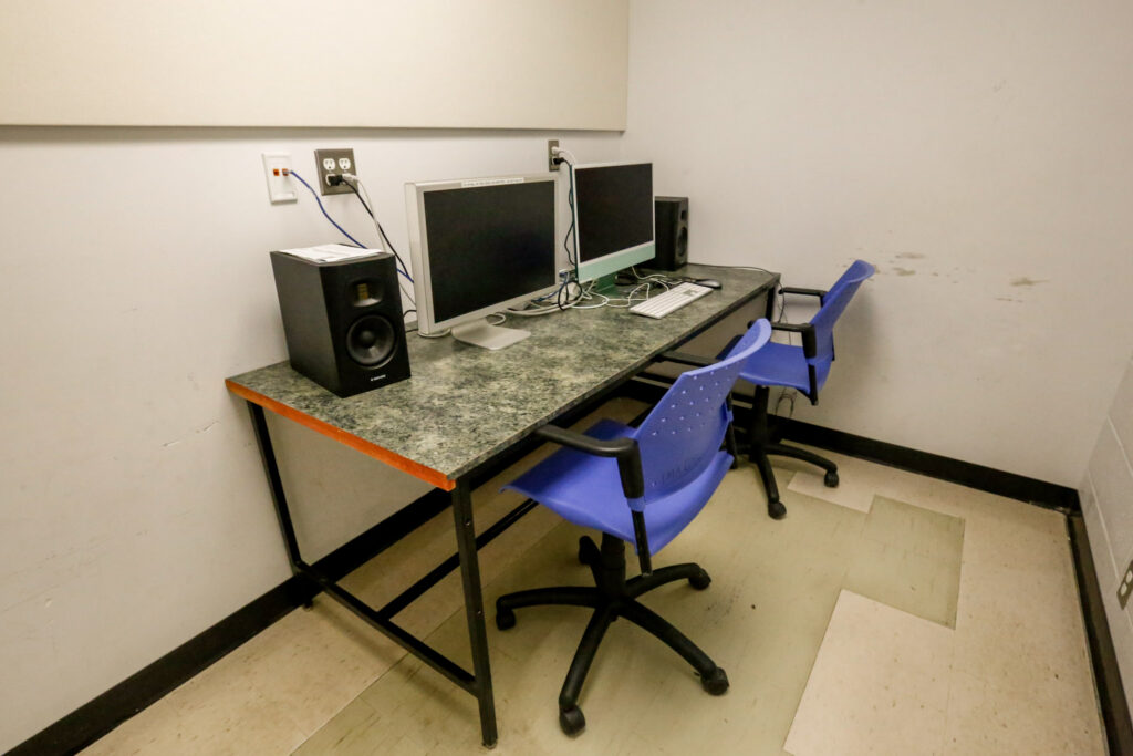An empty computer workstation with two monitors, studio speakers, and two blue office chairs, set in a utilitarian room with plain walls and a tiled floor.