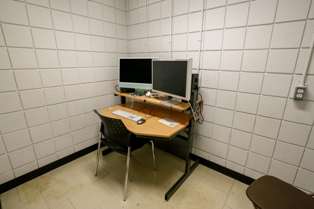 A functional computer workstation in a utilitarian room, featuring two monitors, a keyboard, and a mouse on a wood-top desk, set against a textured white block wall with exposed electrical conduit.