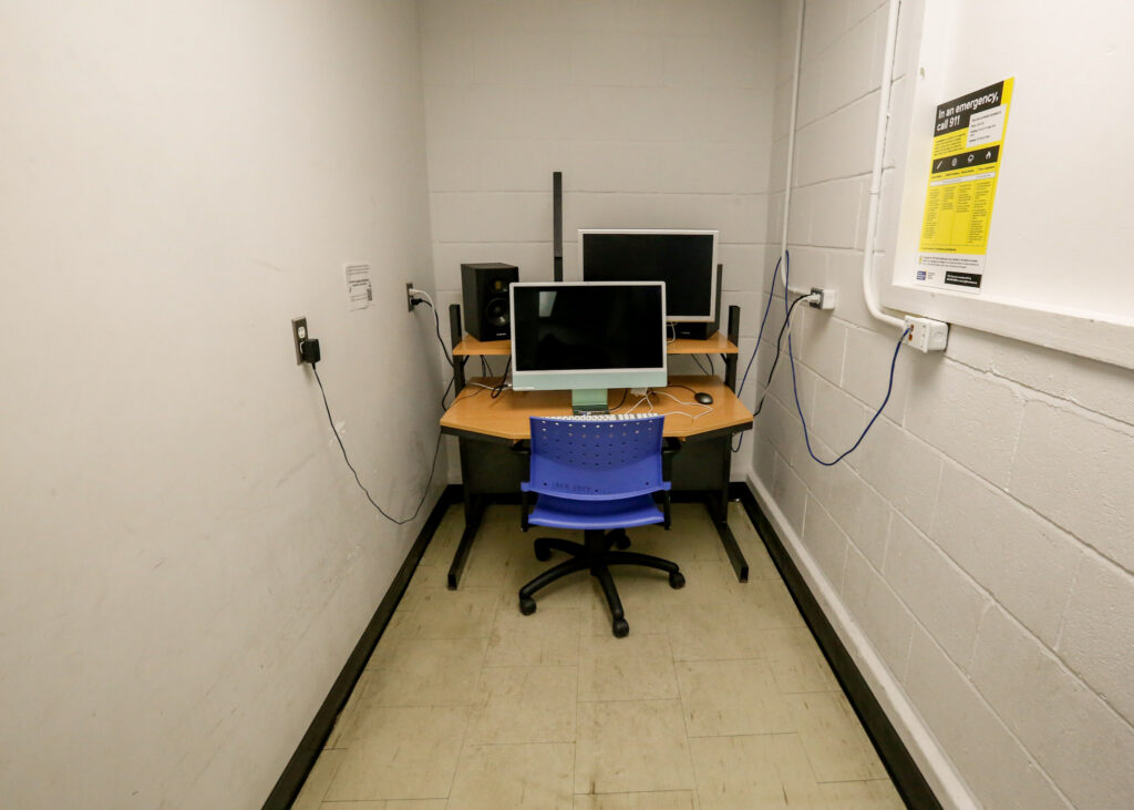 A narrow editing bay or study nook with a corner desk, a light blue iMac, a secondary monitor, and audio speakers, complemented by a blue 'IMA EDIT' chair in a white-walled room with an emergency sign.