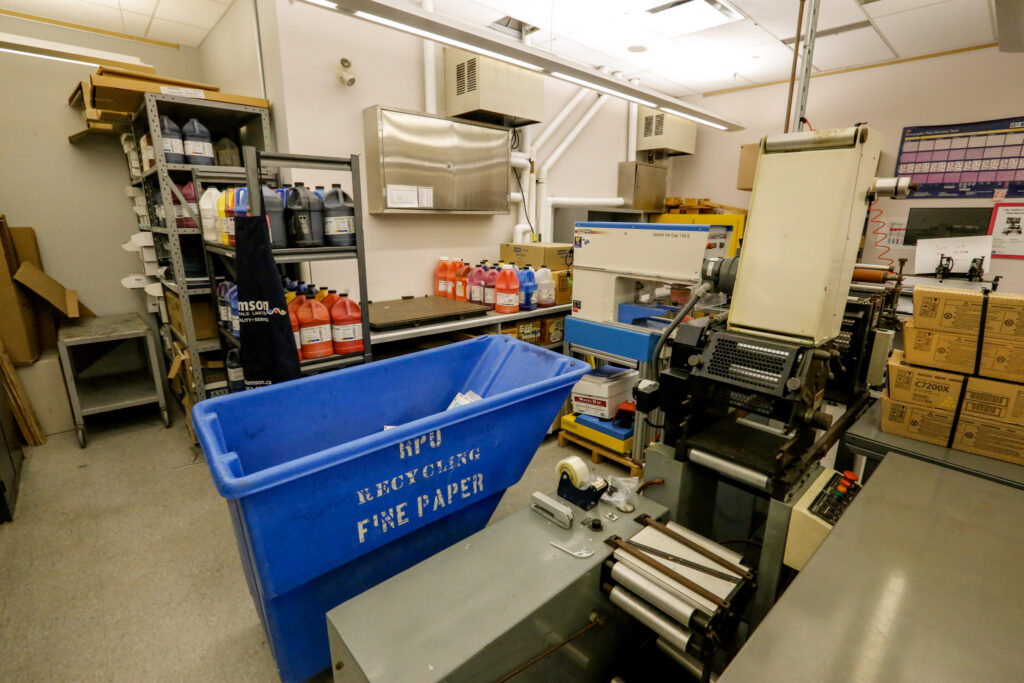 An overview of a busy print shop or industrial facility, showing a large printing press, shelves stocked with ink or chemical jugs, and a prominent blue bin for fine paper recycling.