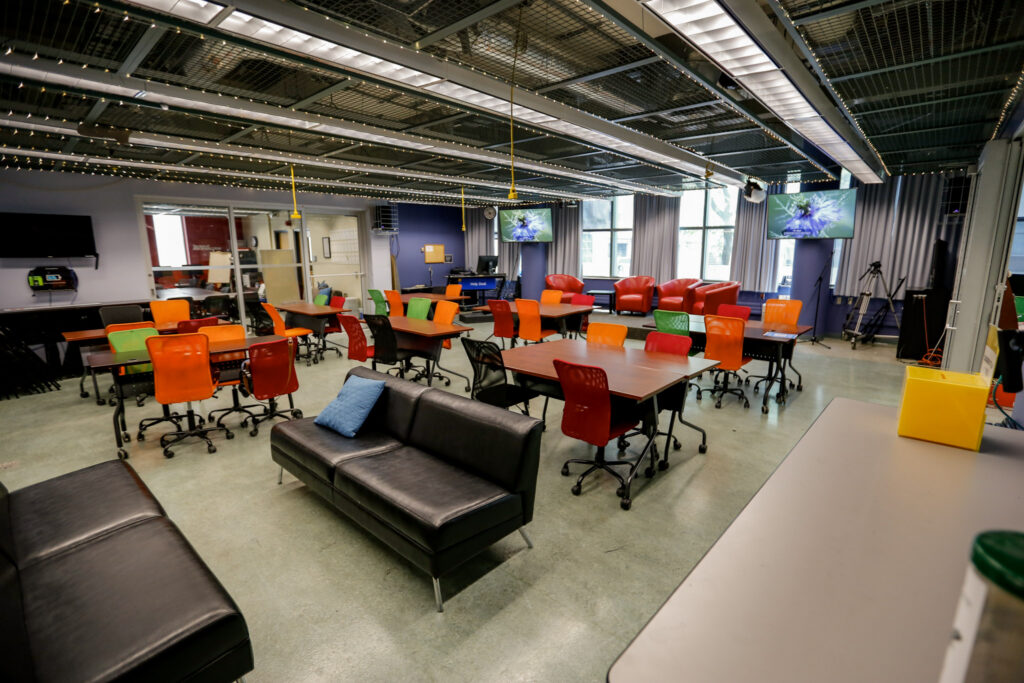 An empty, vibrant collaborative space, possibly a learning lab or flexible classroom, featuring a mix of colorful tables and chairs, comfortable sofas, and lounge seating. Large display screens are mounted on the walls, and decorative fairy lights are strung across the industrial-style ceiling.
